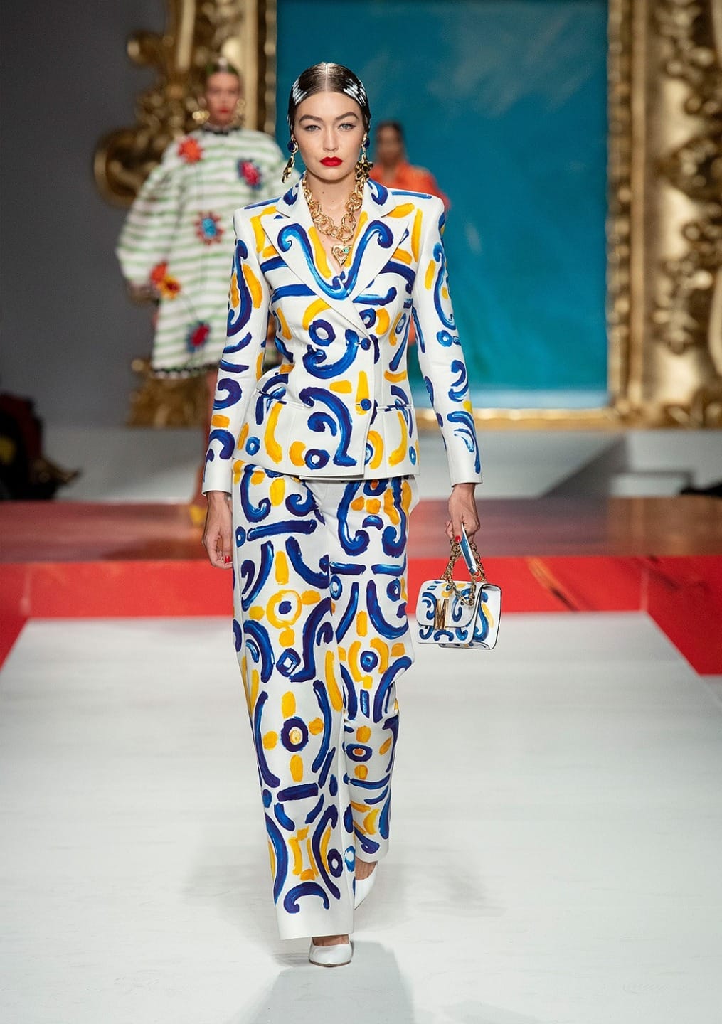 Model walking on fashion runway in vibrant patterned suit with matching handbag, golden accessories, and bold red lipstick.