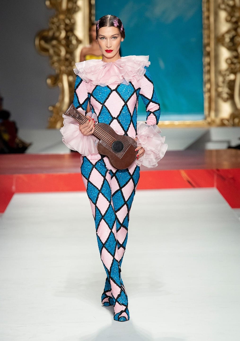 Model walking on runway in colorful harlequin costume with ruffled collar and pink accents, holding small string instrument.