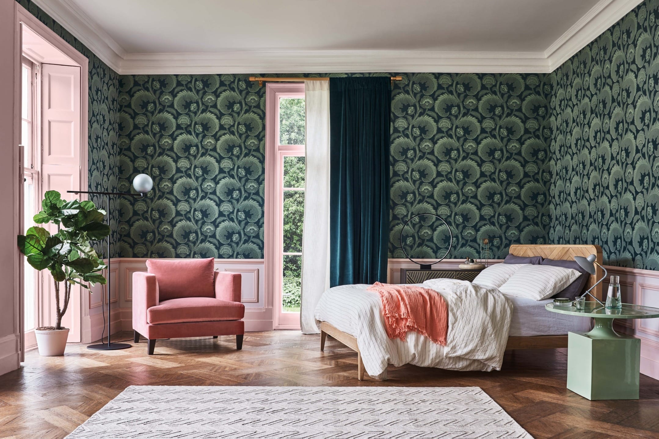 Stylish bedroom with green patterned wallpaper, pink accents, pink armchair, and white bed near large windows.
