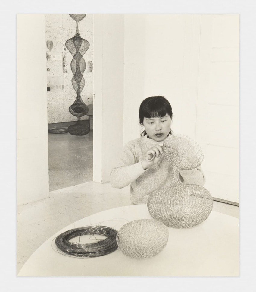 Artist weaving intricate wire sculptures indoors, with several completed pieces and crafting materials on a table nearby.