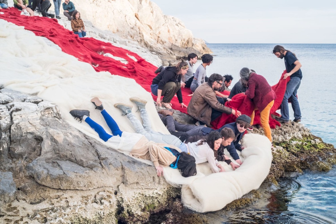 People arranging large red and white fabrics on rocky ocean shore, collaborative outdoor art project in progress.