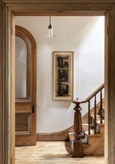 Wooden staircase with ornate railing in a vintage hallway, framed art on white wall, and pendant light above.