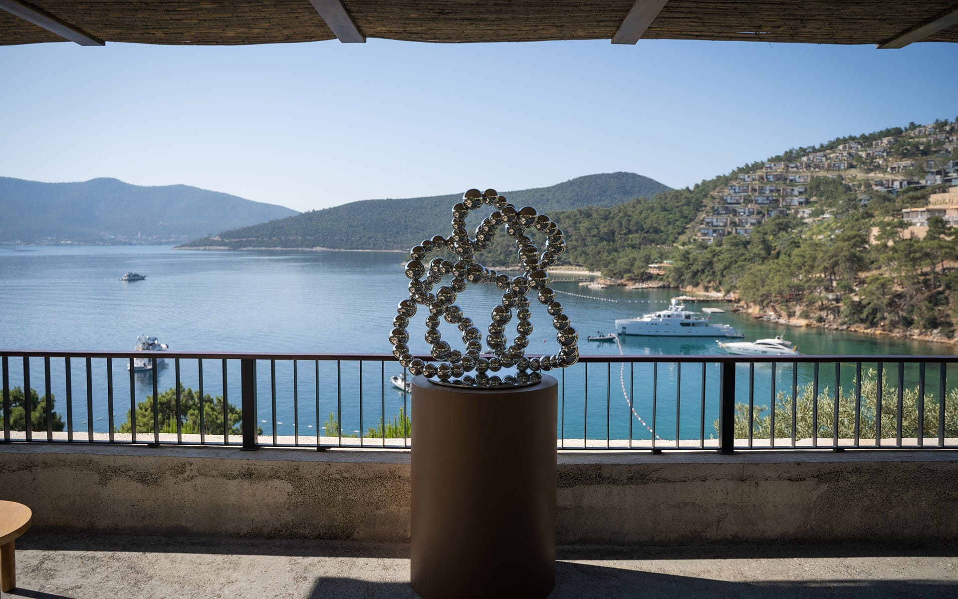 Modern sculpture on terrace overlooking a scenic bay with yachts, surrounded by hills and a clear blue sky in the background.