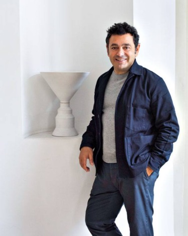 Person smiling in dark jacket standing by a minimalist white sculpture on a white background.