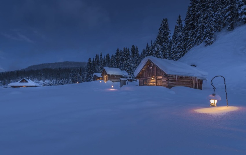 Snow-covered cabins at night with glowing lanterns, surrounded by pine trees in a serene winter landscape.