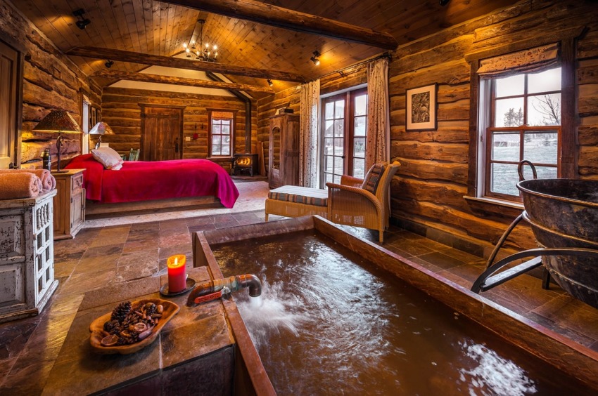 Cozy rustic bedroom with wooden walls, red bedspread, and a stone bathtub by the window, creating a warm atmosphere.