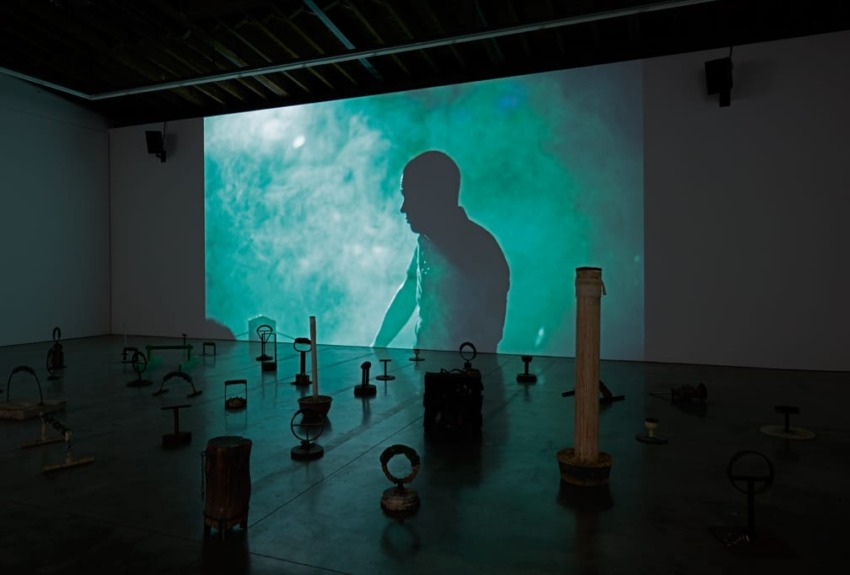Silhouetted figure on green-lit screen in a dark room with scattered abstract sculptures on the floor.