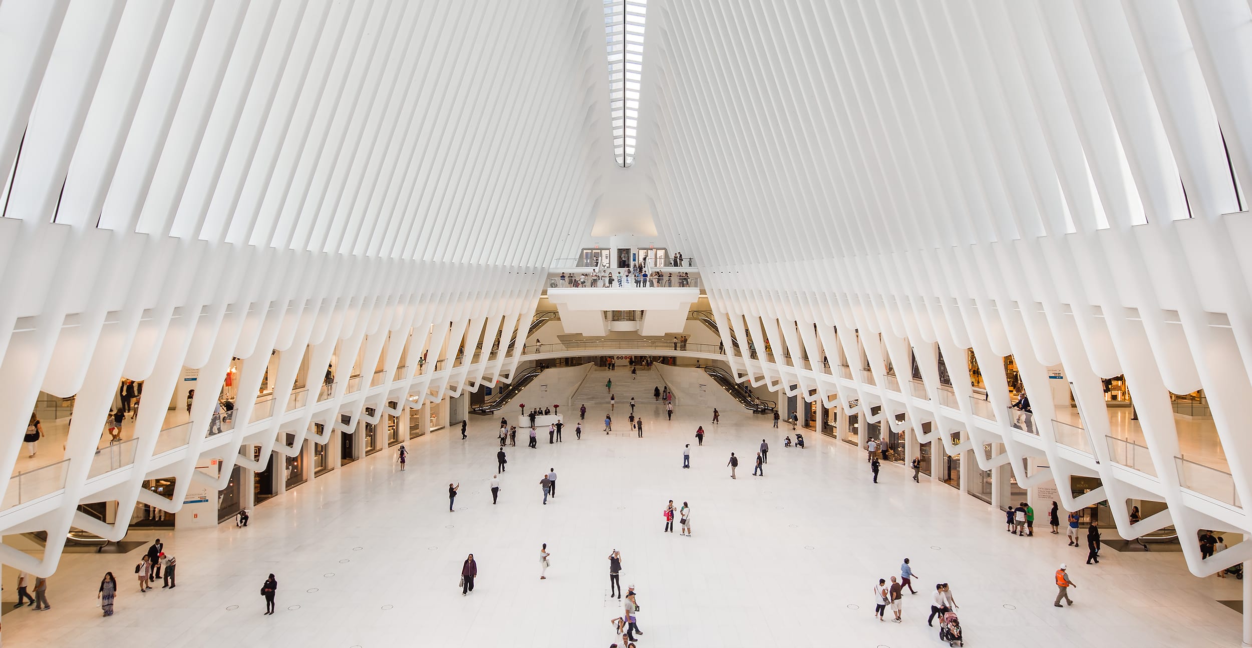 Interior view of a modern architectural space with people walking and white ribbed structures overhead.