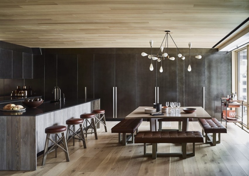 Modern kitchen and dining area with dark cabinets, wooden stools, long table, unique chandelier, and large windows.
