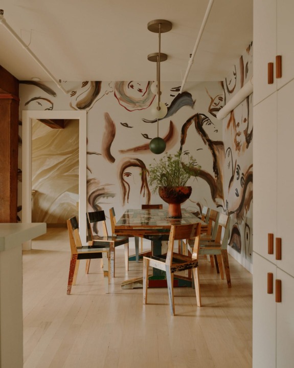 Dining room with abstract face mural, wooden chairs, glass table, and hanging lights.