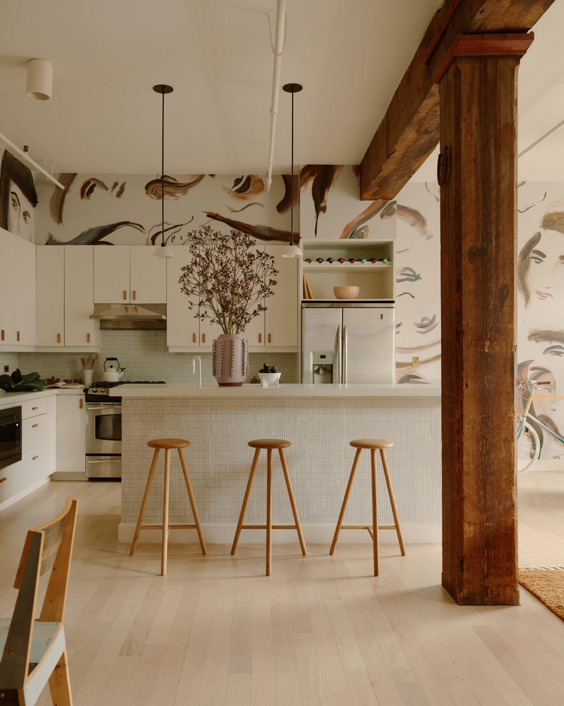 Modern kitchen with white cabinetry, wooden bar stools, large wooden beam, and artistic wall mural in an open living space.