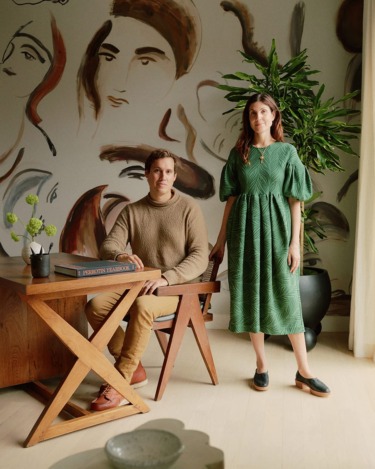A man sitting and a woman standing in a stylish room with artistic wall art and a large potted plant.