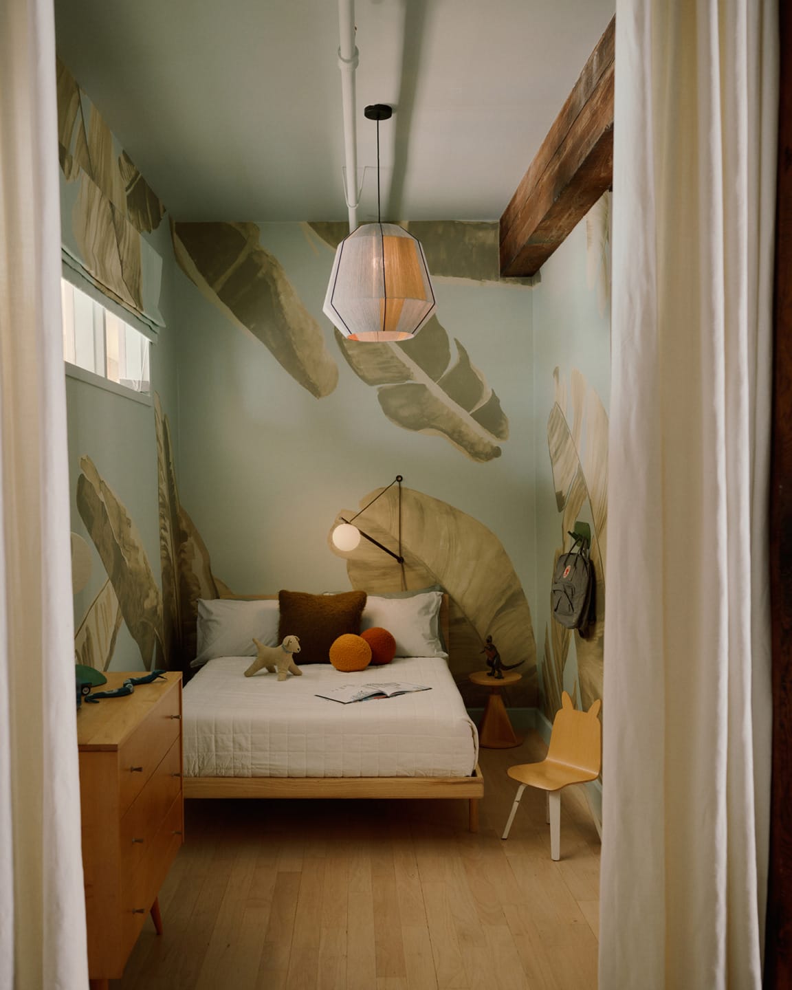 Cozy bedroom with a small bed, decorative pillows, a wooden chair, and a ceiling light fixture, accented by nature-themed wallpaper.