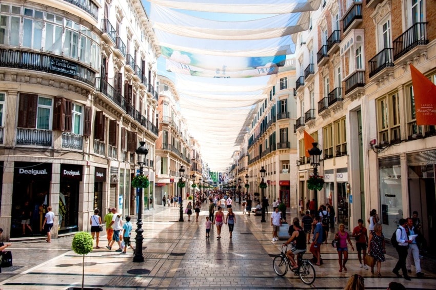 Pedestrian street in a sunny city with people walking, shopping, and cycling under decorative fabric canopies.