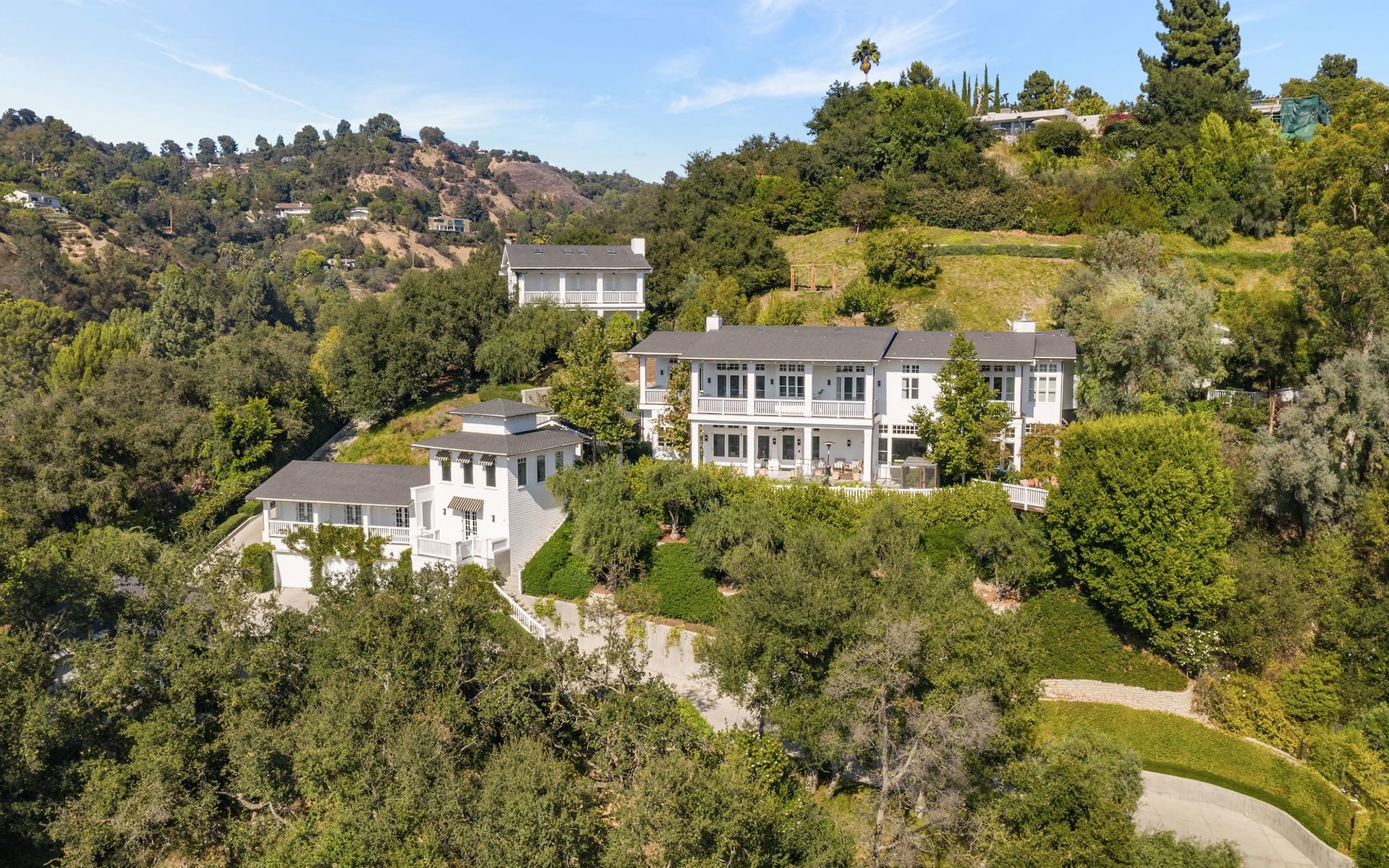 Aerial view of a large hillside house surrounded by trees and greenery under a clear blue sky.