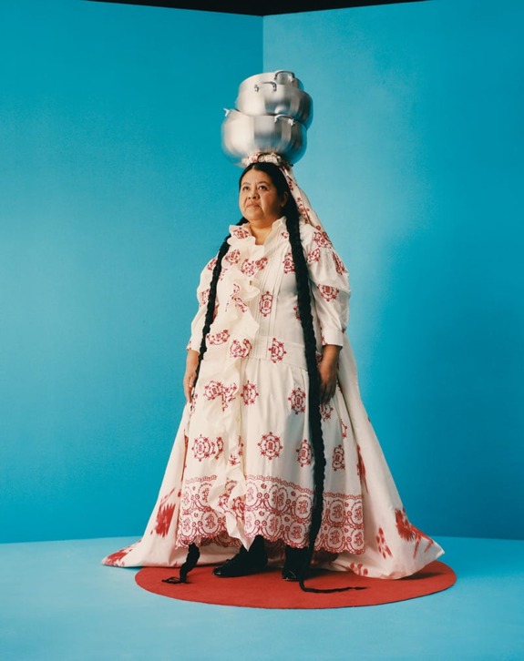 Woman in traditional attire with long braids, balancing silver pots on head, standing on a red circle against blue background.