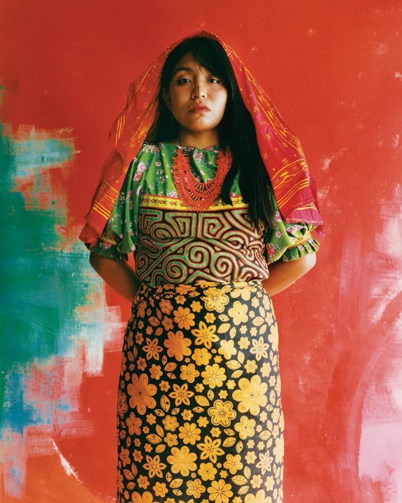Woman in colorful traditional attire with patterned top, floral skirt, and red textured veil against vibrant red background.