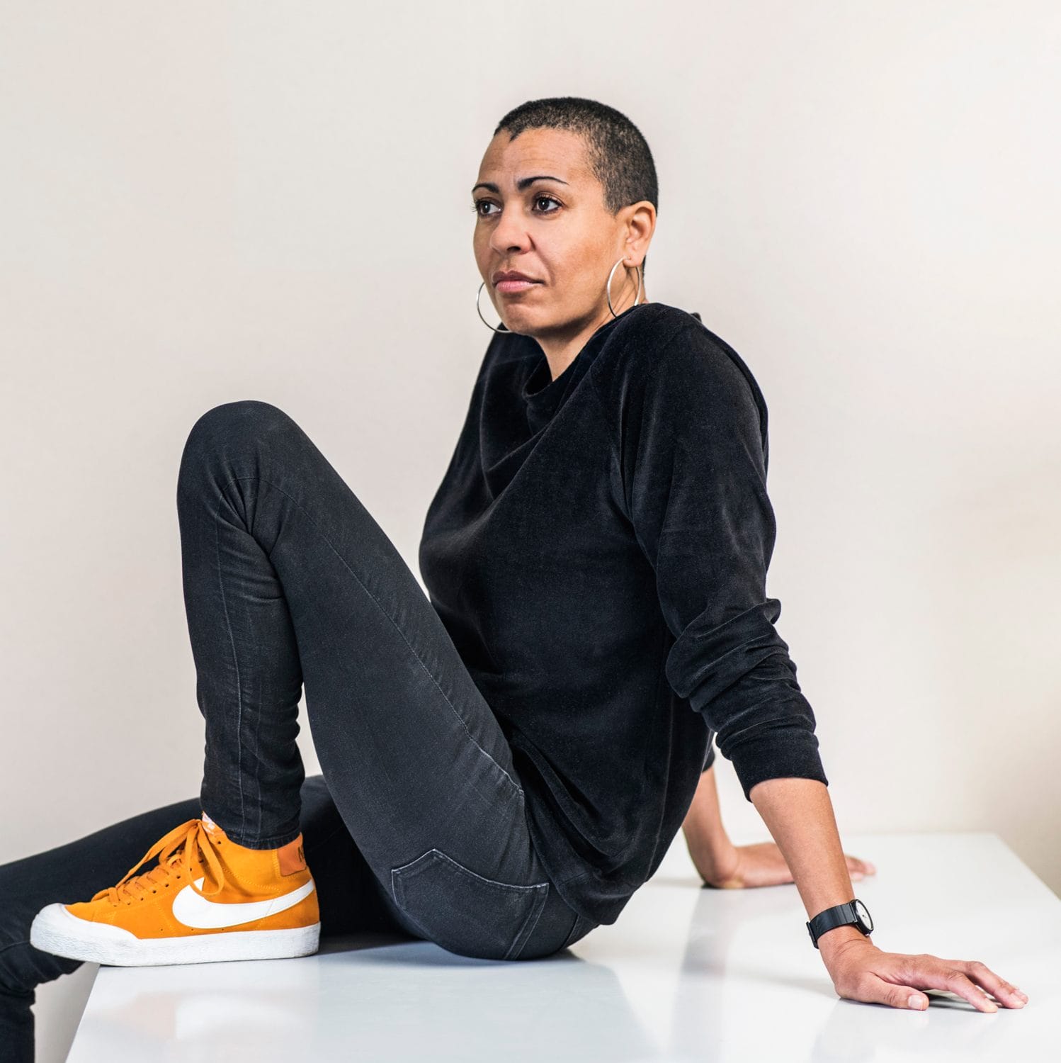 Person sitting on a white surface, wearing a black outfit and bright orange sneakers, looking to the side.