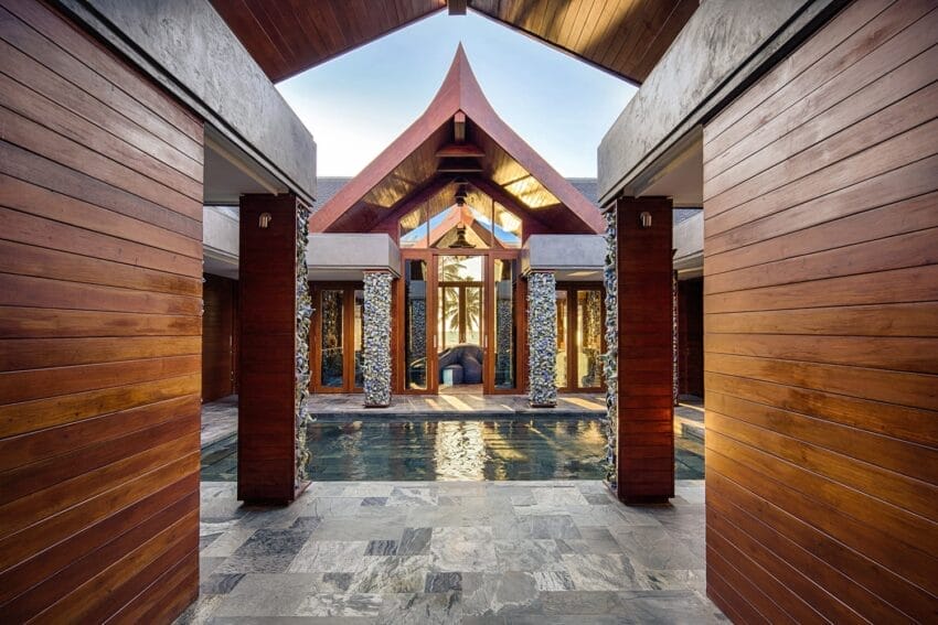 Luxury villa entrance with wooden panels, stone accents, and water feature, leading to high ceiling and large windows.