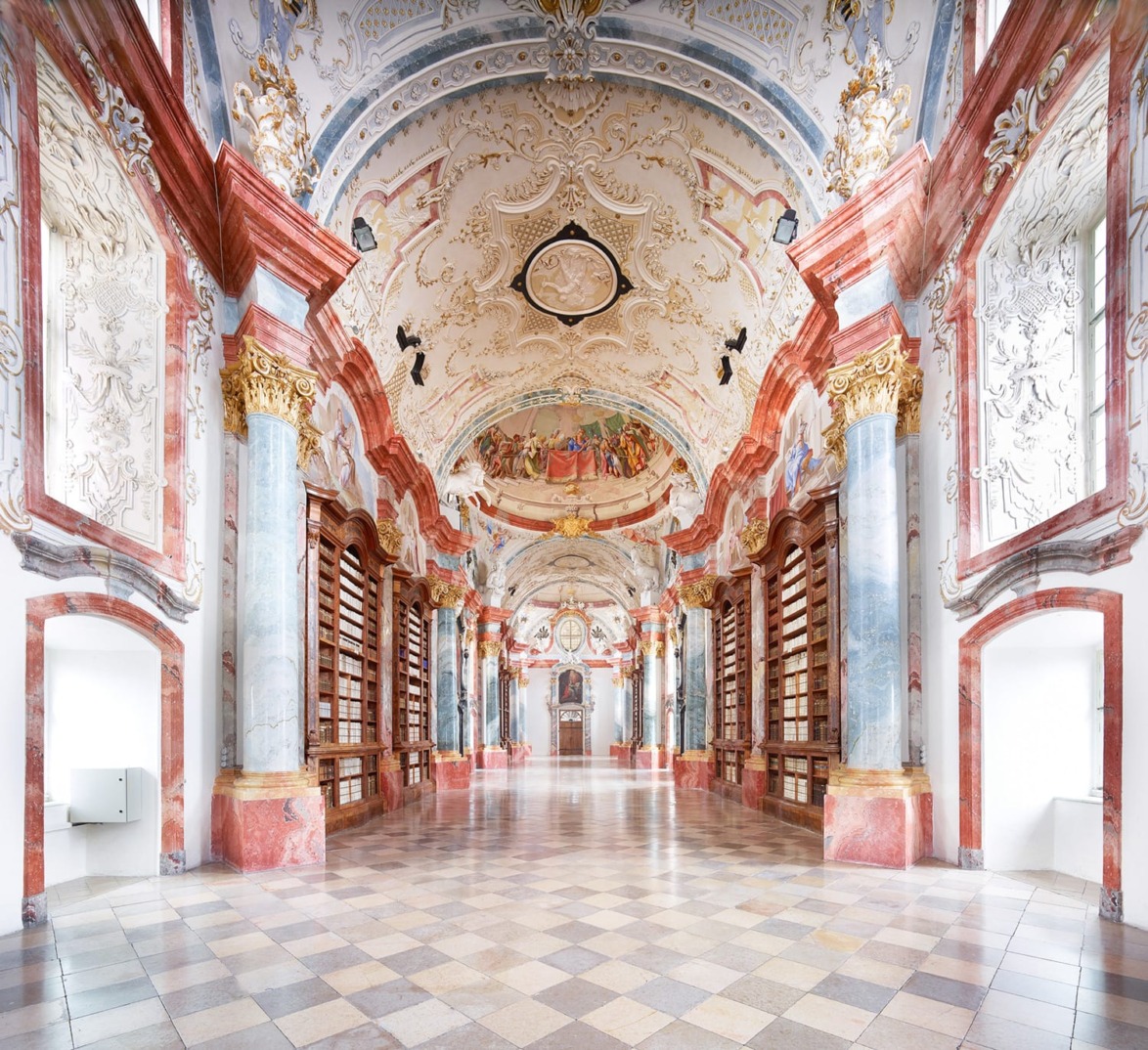 Ornate library interior with tall marble columns, intricate ceiling artwork, and rows of bookshelves lining the walls.