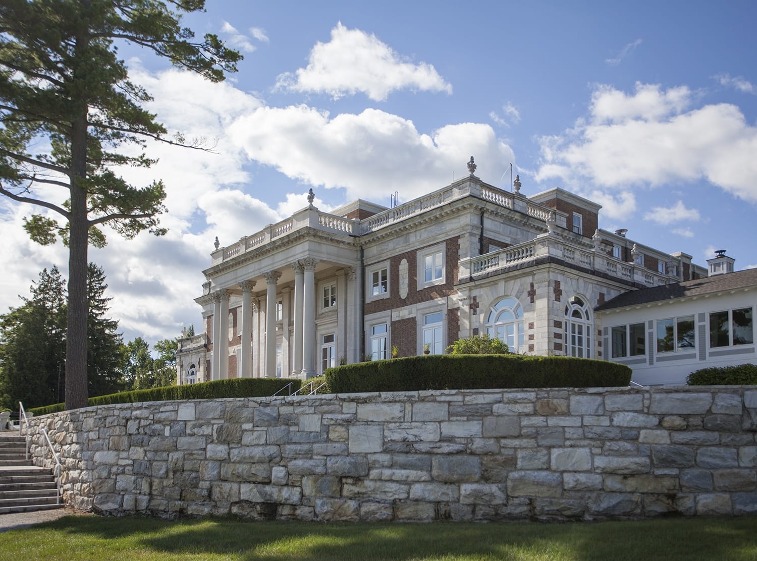 Large historic mansion with classical architecture, surrounded by stone wall and trees under a blue sky with scattered clouds.