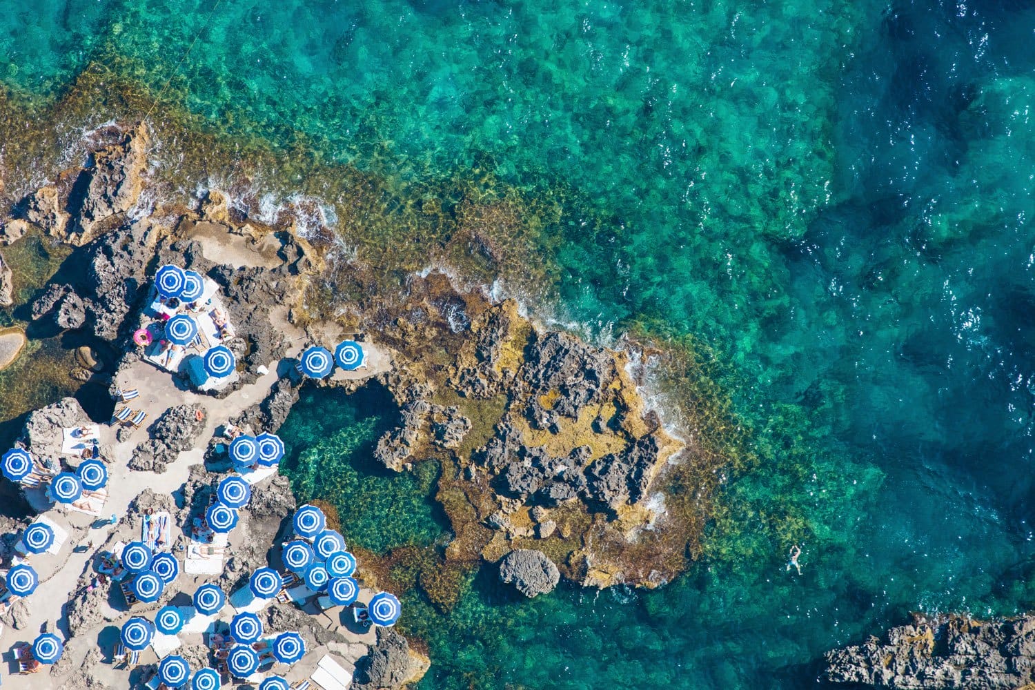 Aerial view of rocky coastline with turquoise waters and blue-and-white striped umbrellas on a beach.
