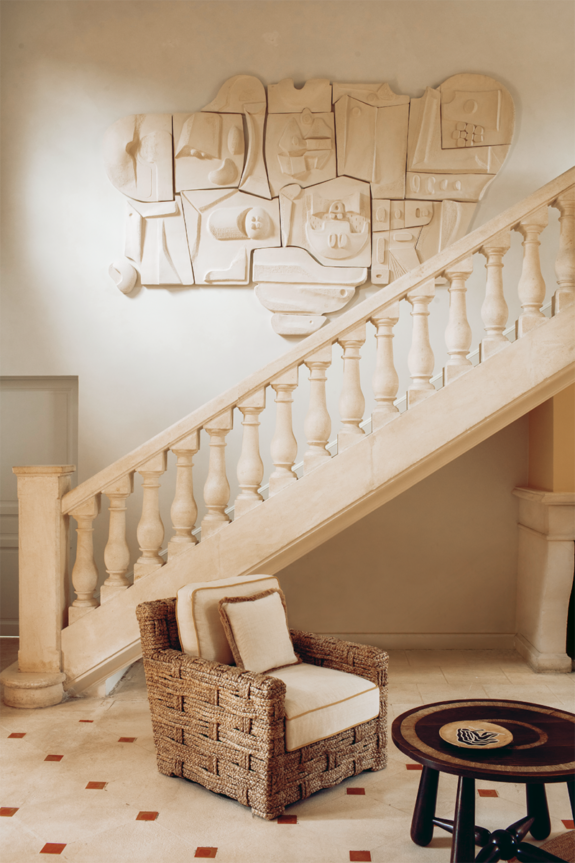 Elegant staircase with intricate wall art above, wicker armchair, and small round table on a tiled floor.