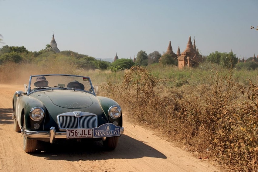 Classic convertible car driving on a dirt road with ancient temples in the background on a sunny day.