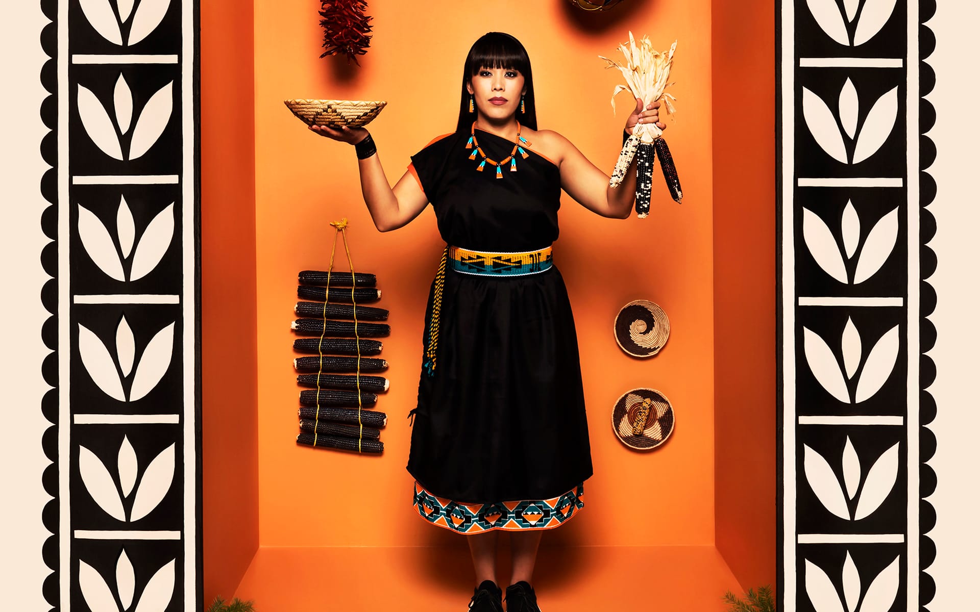 Person in traditional attire holding a bowl and corn, standing against an orange background with decorative elements.