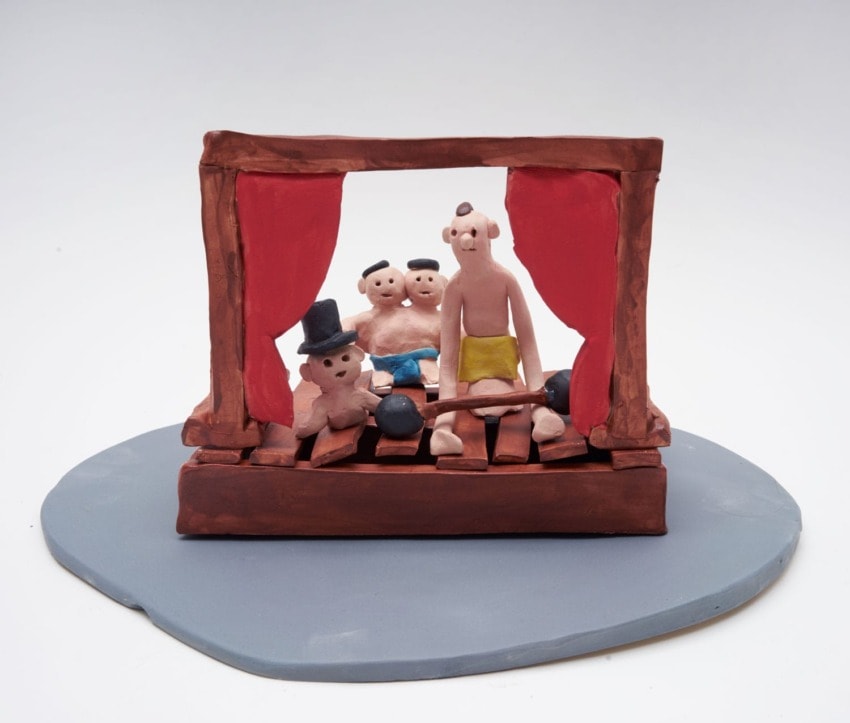 Clay figurines performing on a stage with red curtains, depicting a whimsical circus scene with a strongman and spectators.