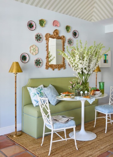 Elegant dining area with green bench, round table, floral arrangement, gold lamps, and decorative plates on wall.