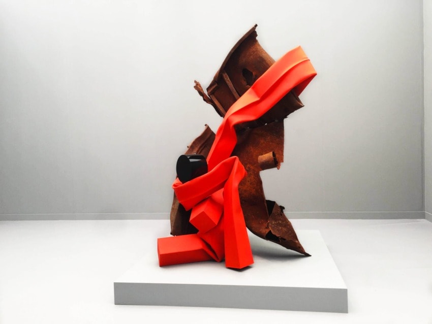 Sculpture with rusted metal and vibrant red ribbon-like shapes on a white platform against a neutral background.