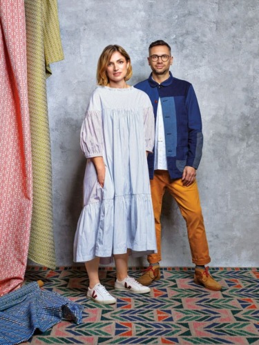 Two people standing on a patterned rug, wearing stylish clothing, with textured fabric hanging in the background.