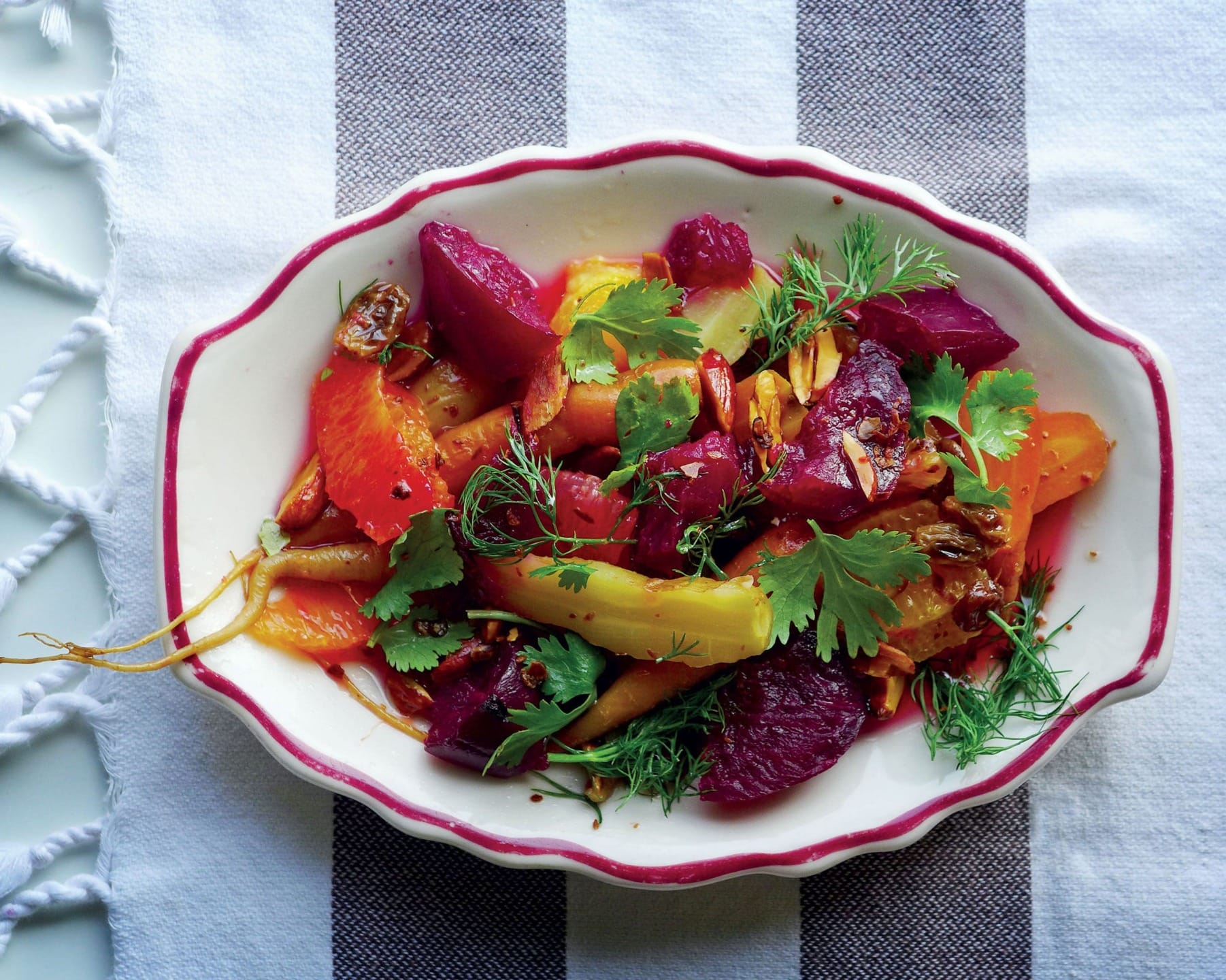 Colorful roasted beet salad with oranges, herbs, and seeds in a white dish on a striped tablecloth.