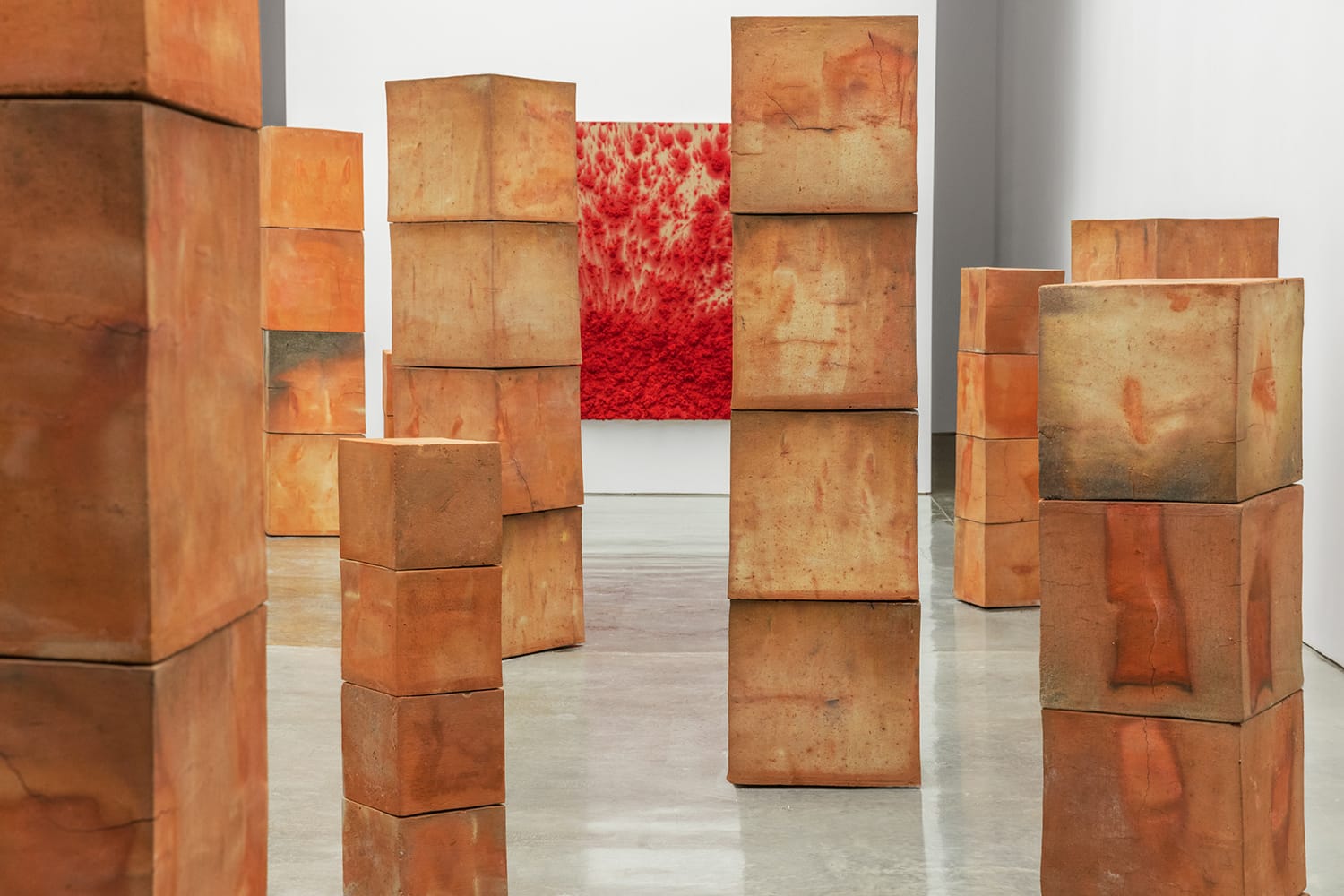 Abstract art installation with tall rectangular terracotta sculptures, red textured painting in the background, polished floor.