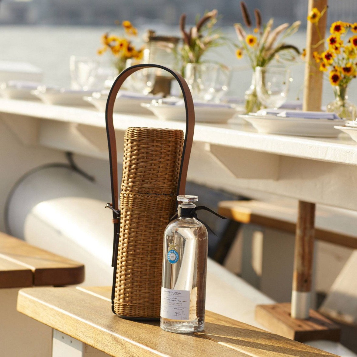 Picnic table on a boat with a wicker basket and a bottle of water, surrounded by plates, glasses, and flowers.