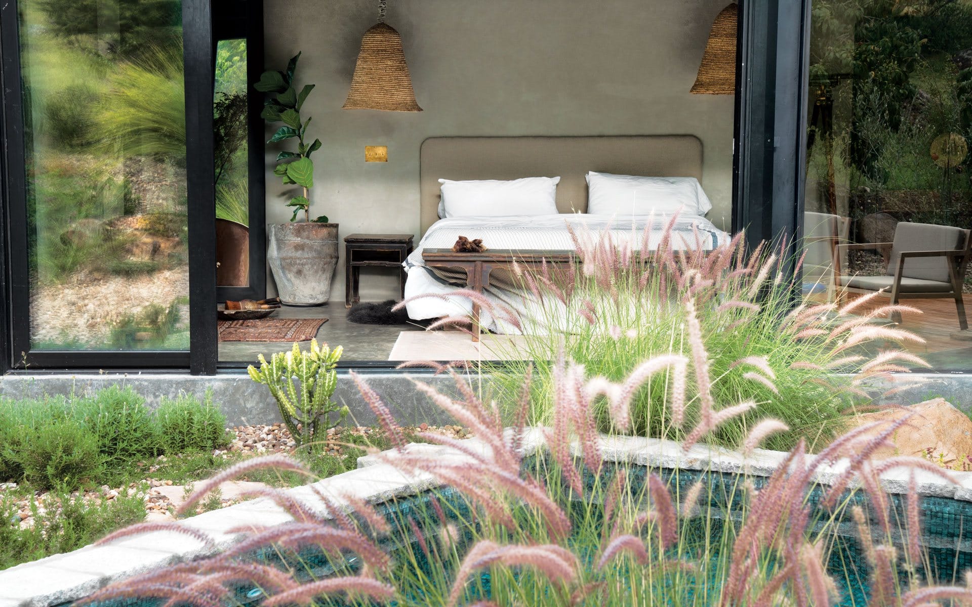 Modern bedroom with glass wall, overlooking a garden and pool, featuring a cozy bed and natural decor elements.
