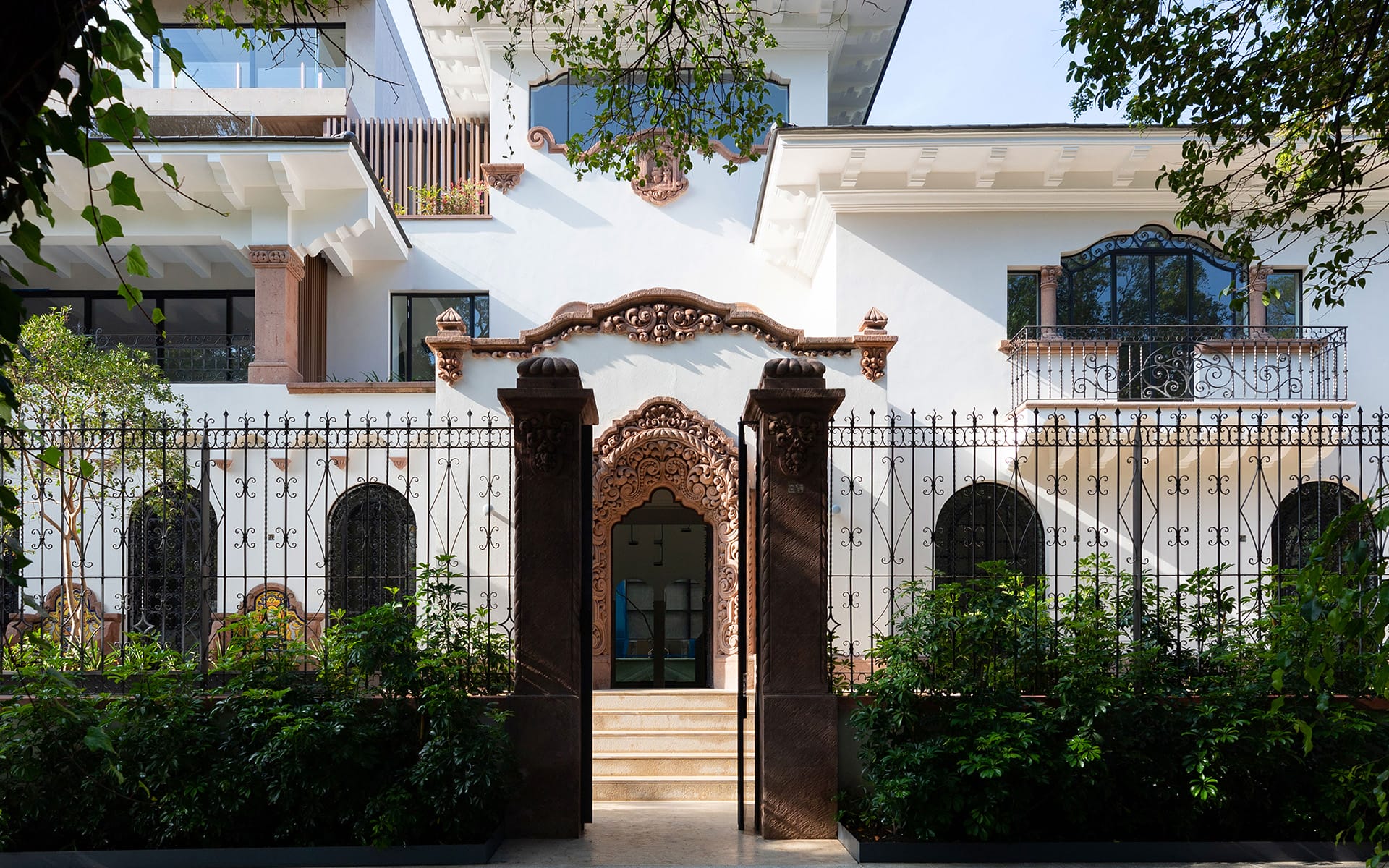Ornate two-story white building with decorative iron fence, arched windows, and lush greenery at the entrance.