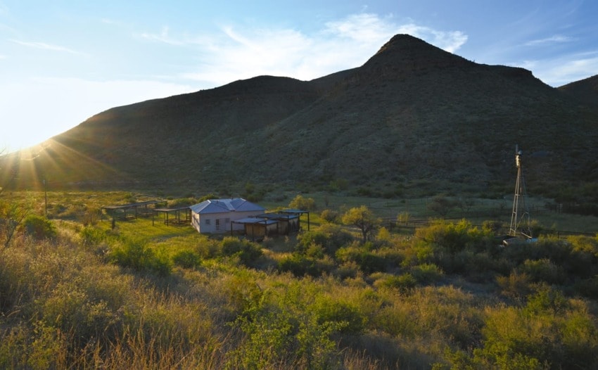 Rural landscape with a house, windmill, and mountains under a clear sky at sunset.