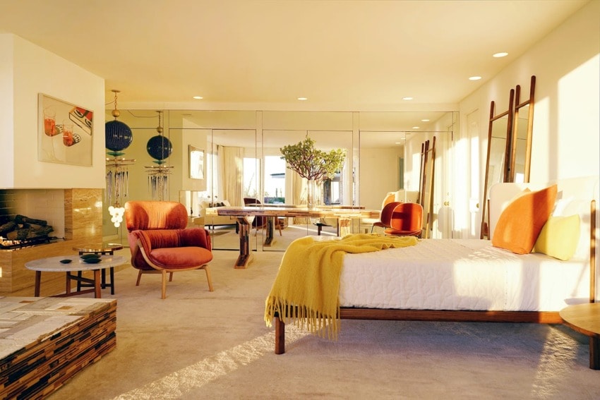 Mid-century modern bedroom with orange accents, cozy seating, and natural light through large windows and mirrors.