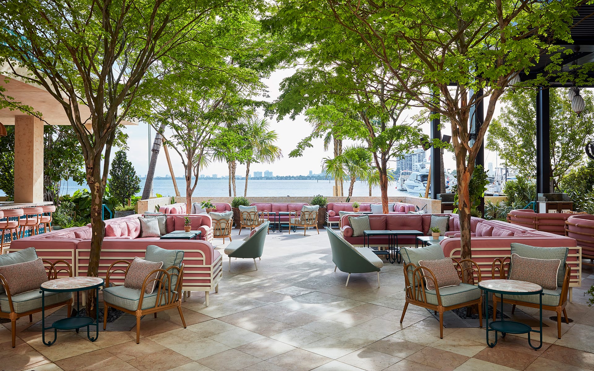 Outdoor restaurant patio with pink seating, tables, and lush green trees overlooking a waterfront view.