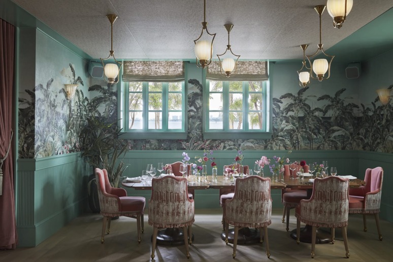 Elegant dining room with green walls, floral wallpaper, ornate chairs, a long table set for a meal, and stylish hanging lights.