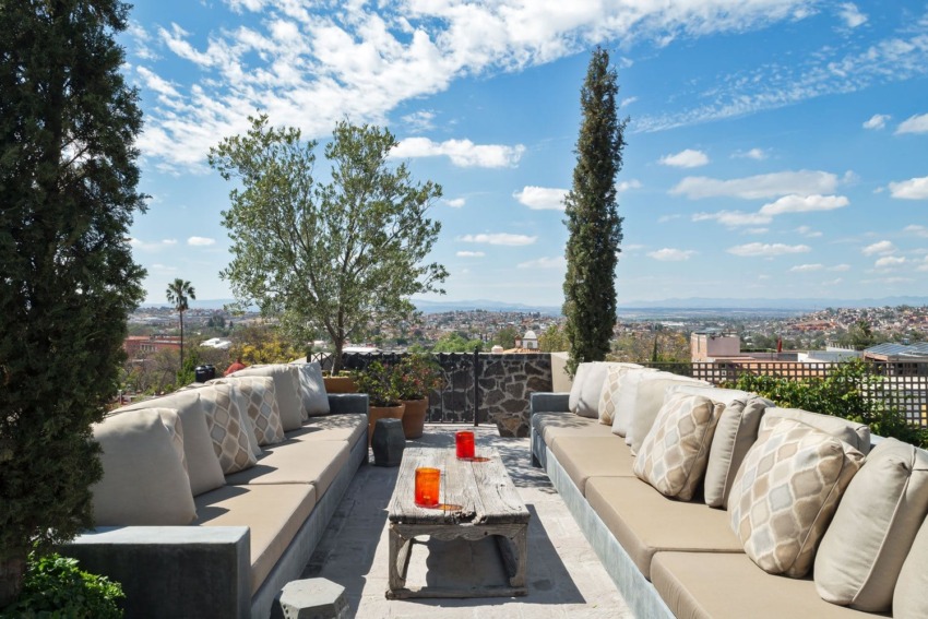 Outdoor patio with beige couches, stone table, and scenic view of city and sky with clouds and trees.