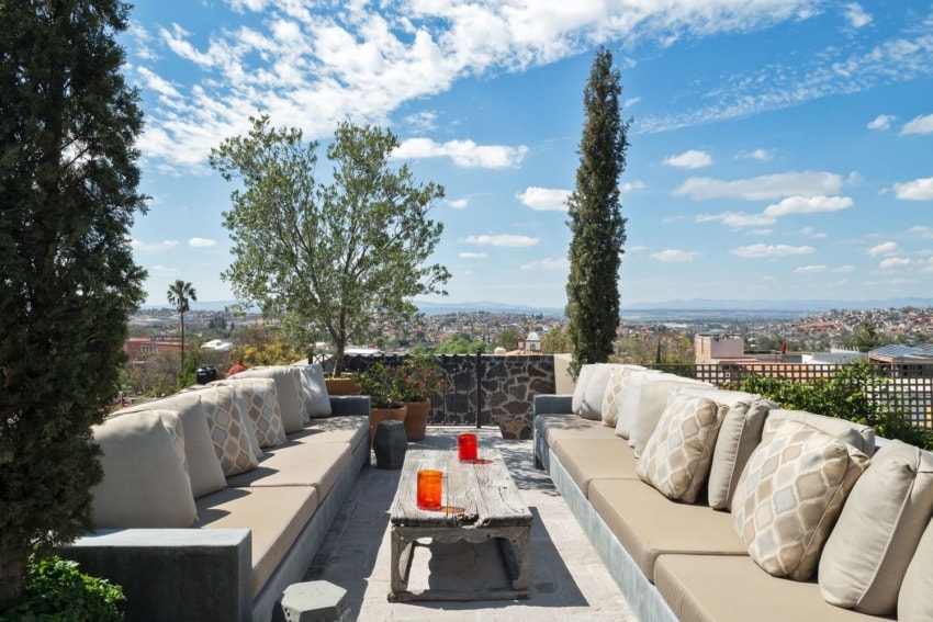 Outdoor patio with beige couches, stone table, and scenic view of city and sky with clouds and trees.