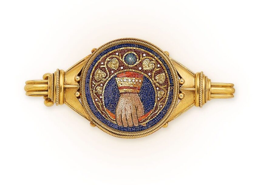 Ancient gold brooch with intricate mosaic design featuring a hand surrounded by decorative patterns on a deep blue background.