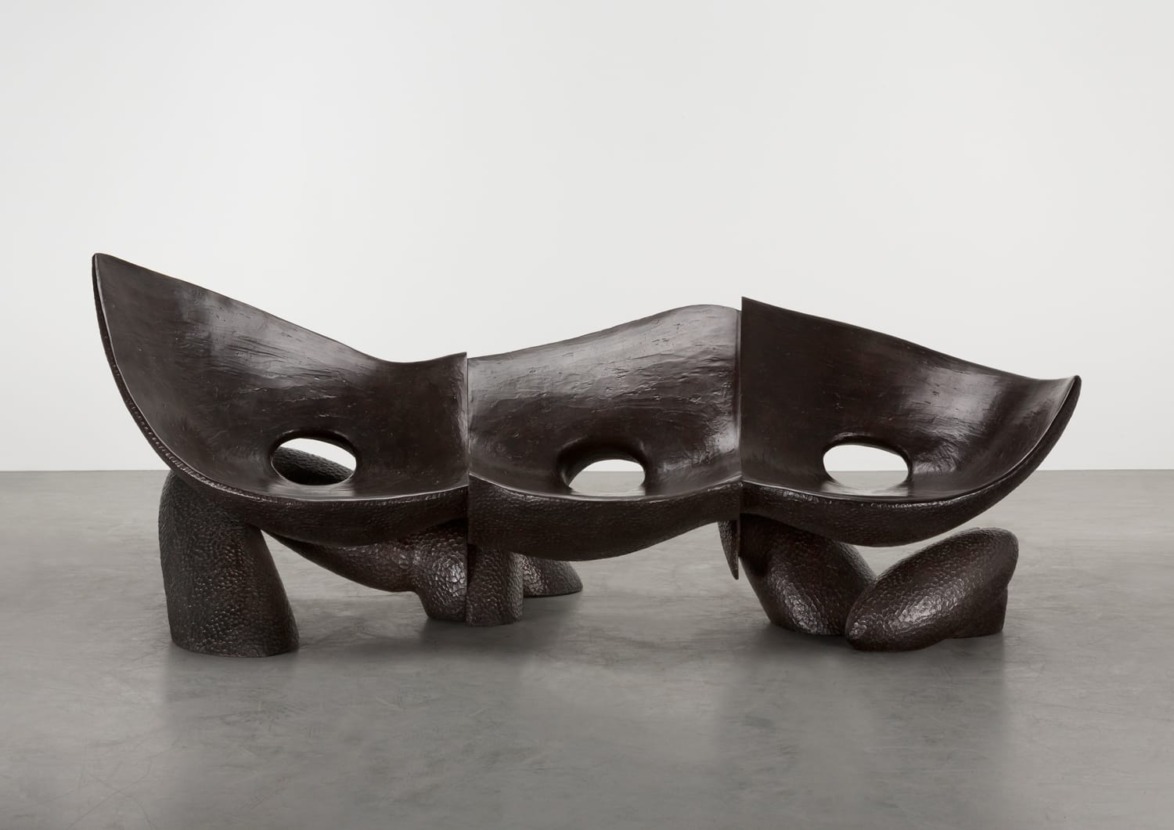 Abstract bronze sculpture with three interconnected curved seats and openings, displayed on a gray floor and background.