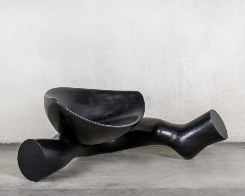 Sleek modern black sculpture with intertwined shapes resembling a seat on a concrete floor against a plain wall.