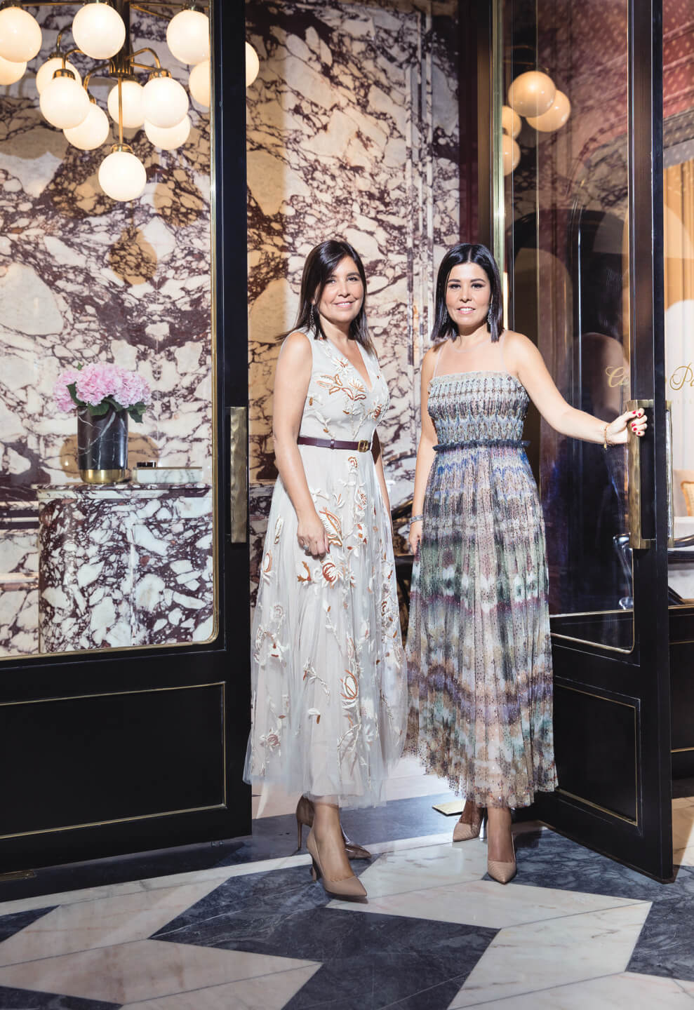 Two women in elegant dresses stand in an ornate entrance with marble walls and a stylish backdrop.