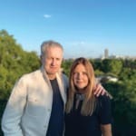 Man and woman standing together outside with a cityscape and trees in the background on a sunny day.