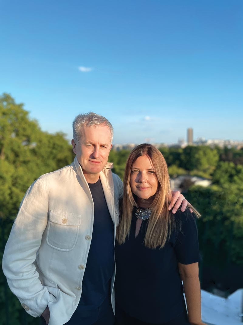 Man and woman standing together outside with a cityscape and trees in the background on a sunny day.